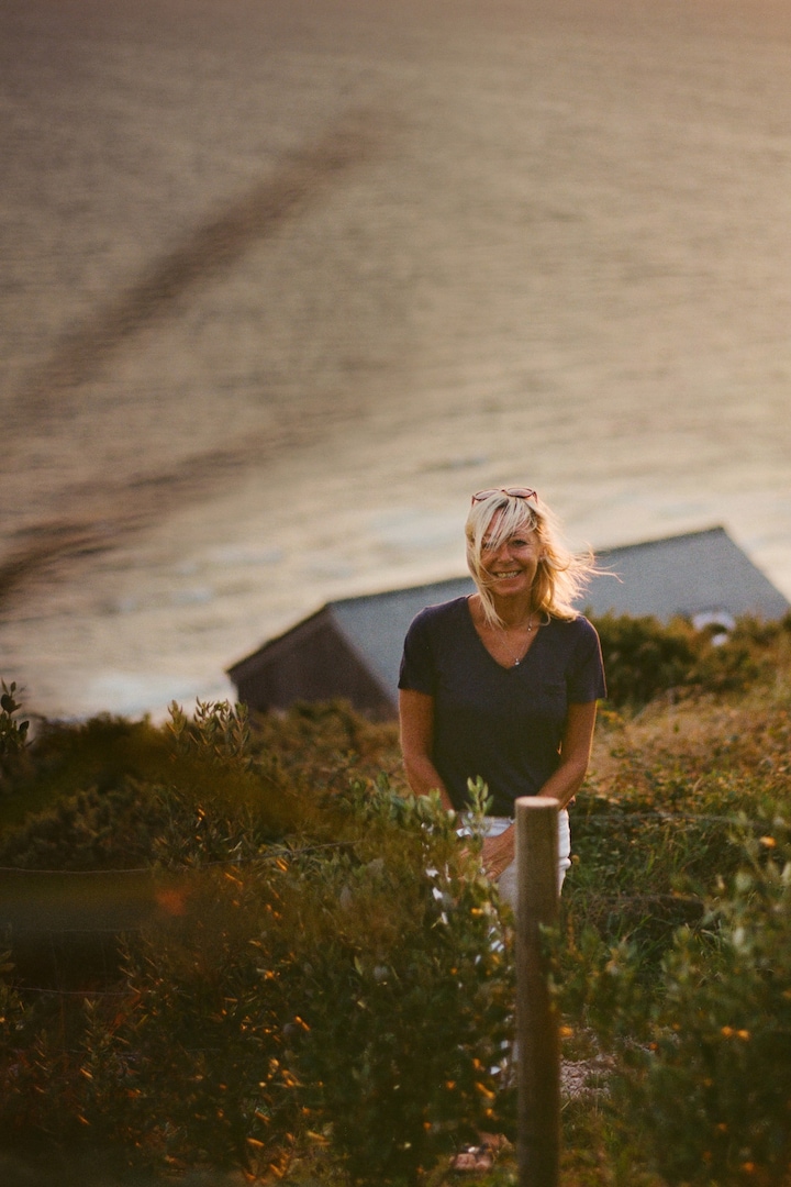 Debout dans un champ, une hôte sourit chaleureusement. L'océan et le toit de la maison se situent derrière elle, en arrière-plan.