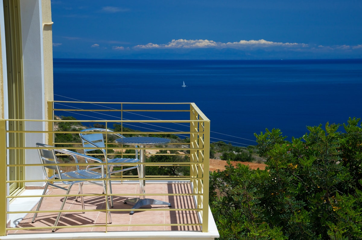 A balcony features two metal chairs and a small table, offering a clear view of the expansive blue sea. The horizon is marked by distant clouds, while lush greenery frames the scene, creating a serene outdoor space.
