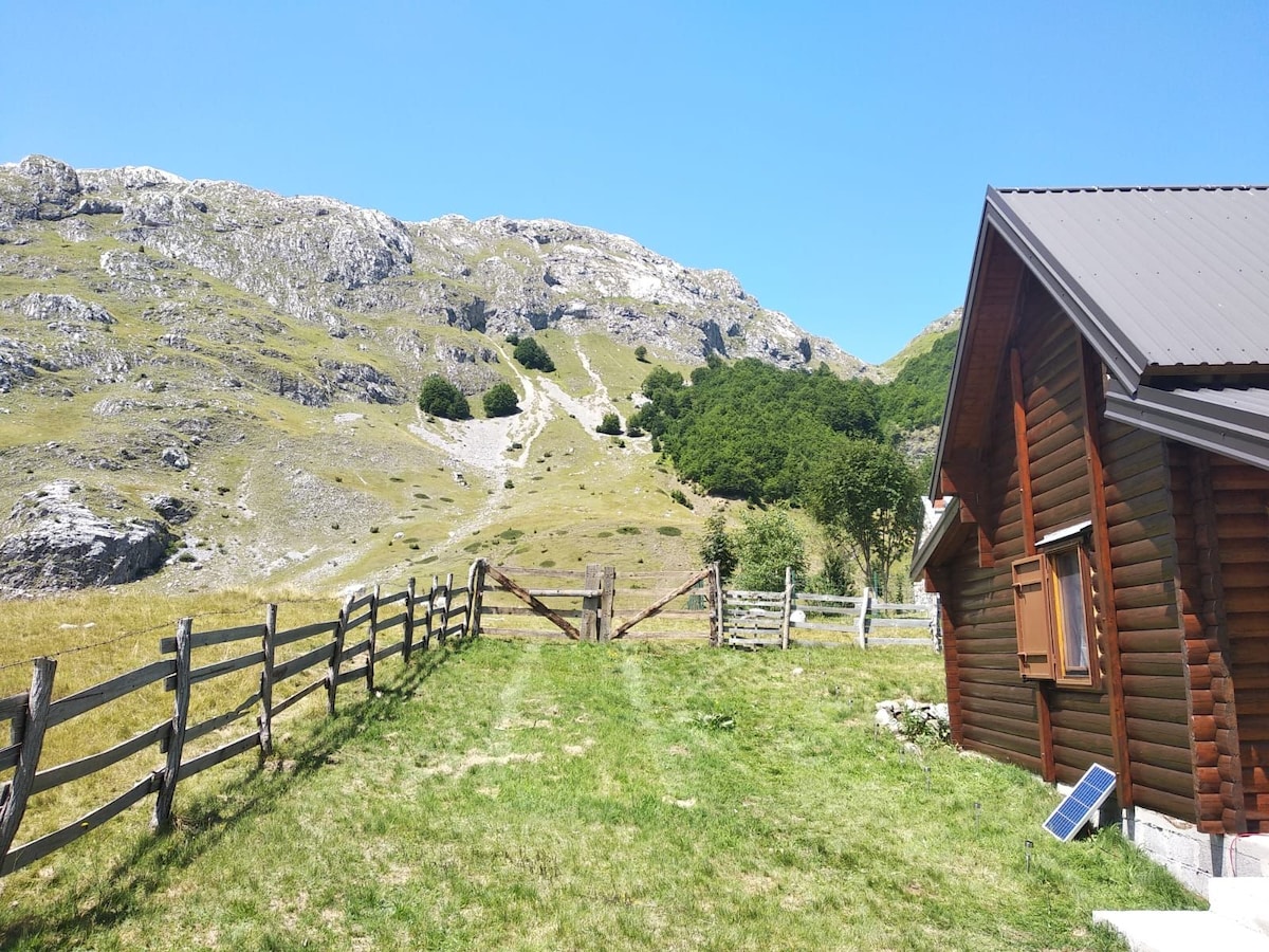 A wooden cottage is depicted beside a grassy area, with a fence leading to rolling hills and rocky mountain peaks in the background. Clear blue skies are visible above, highlighting the natural landscape and serene atmosphere surrounding the cottage.