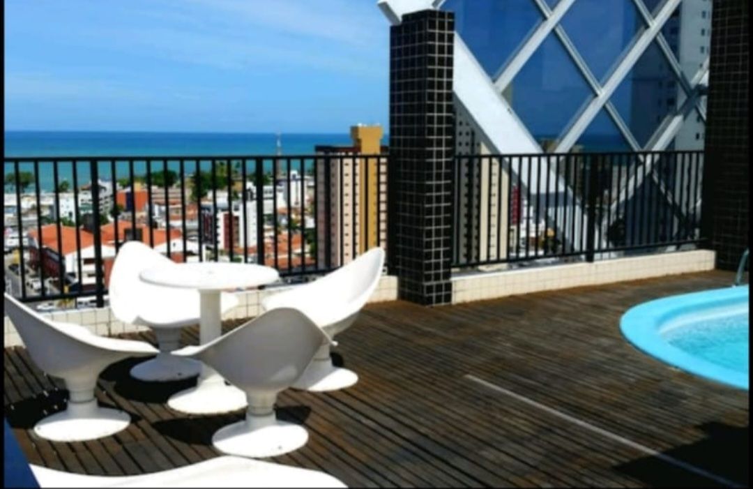 A rooftop terrace features a seating area with white chairs and a round table, set against a backdrop of the ocean and city skyline. A small hot tub is visible beside the seating area, and wood decking adds warmth to the space.