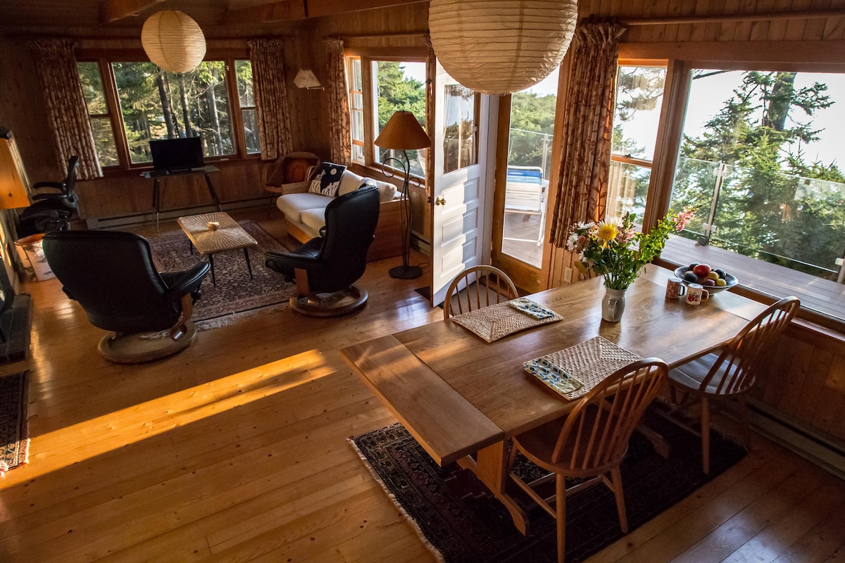 A cozy great room features a dining table with wooden chairs, illuminated by natural light. Two comfortable chairs are positioned around a small table, while large windows offer views of the outdoors. Floral arrangements and fruit add color to the inviting space.