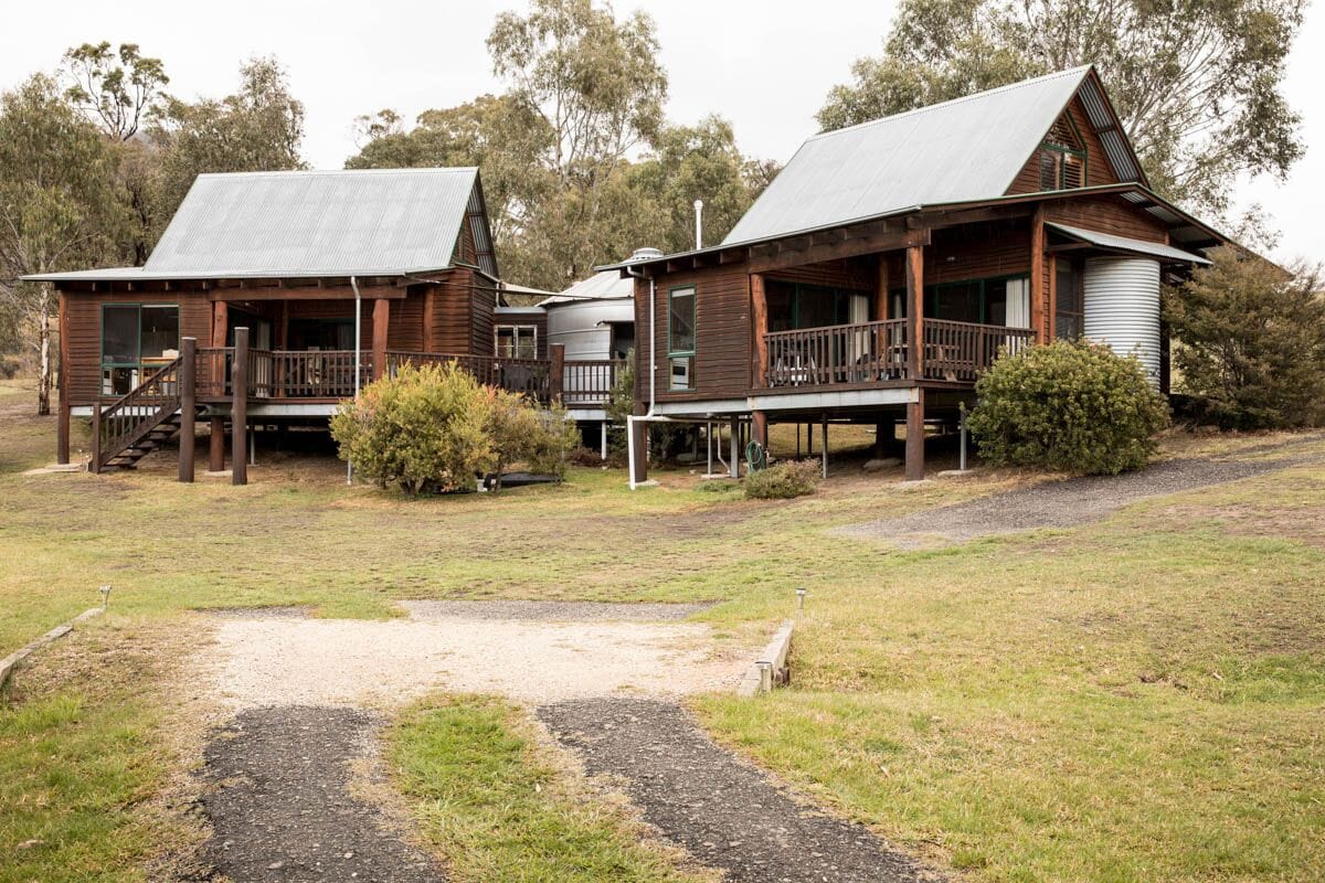 Two architect-designed eco-friendly cabins are surrounded by a spacious yard, each with separate entrances and decks. Natural timber features prominently in their construction, and lush greenery lines the pathways leading to both cabins.