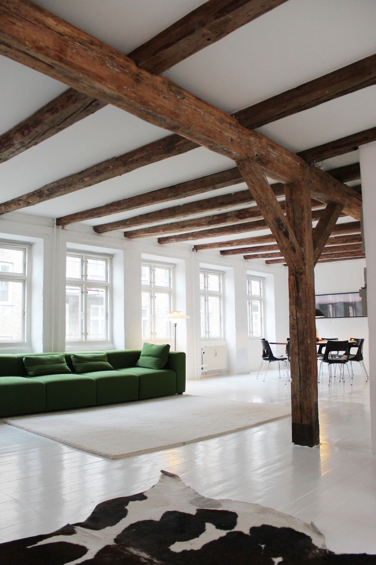 A spacious living area features a green sectional sofa and a large white rug. Exposed wooden beams add character to the ceiling, while multiple tall windows provide ample natural light. A dining area with black chairs is visible in the background.