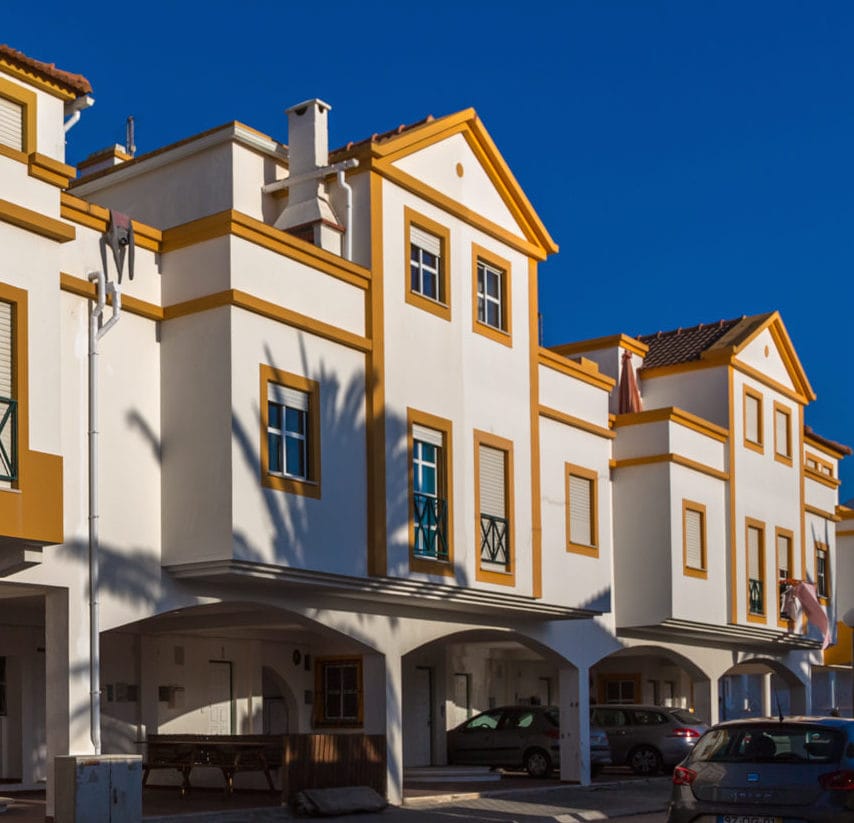 A two-story apartment building features a clean white facade with vibrant yellow accents. The structure includes several large windows and balconies, allowing for ample natural light. The ground level is partially covered, providing shaded seating areas. Palm trees create a tropical ambiance in the background.