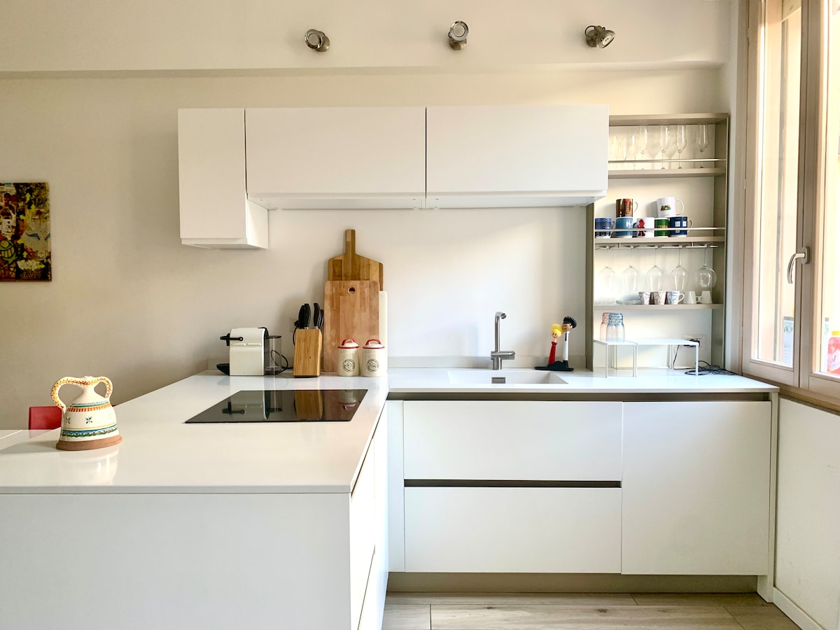 A modern kitchen is depicted, featuring sleek white cabinetry and an island with a built-in induction cooktop. Glassware and utensils are organized on open shelves, while natural light streams in from a nearby window, illuminating the minimalist design.