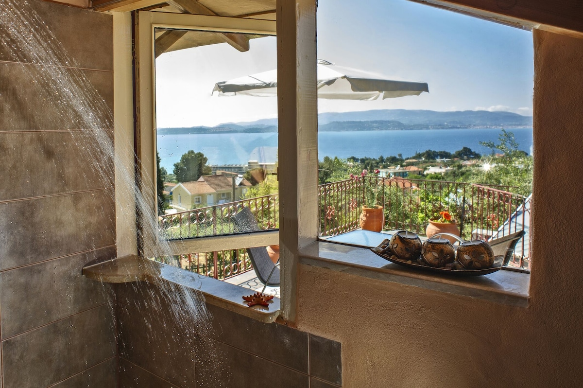 A spacious bathroom features a window that frames a scenic view of the sea and surrounding landscape. Natural light floods the area, reflecting off the stone tiles. A water feature runs down the wall, enhancing the ambiance of relaxation.
