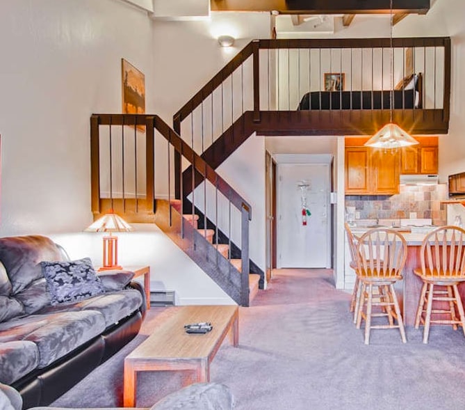 An interior view of a loft condo showcases a cozy living area with a dark sofa and a wooden coffee table. A wooden staircase leads to the loft, where a queen bed can be seen. The kitchenette and dining area with four chairs are visible in the background.