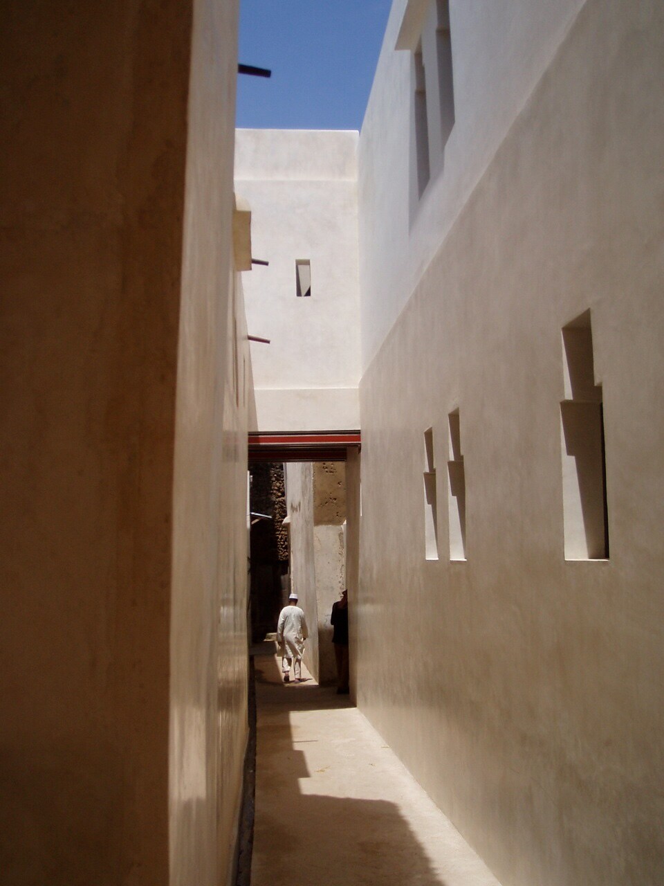 A narrow corridor is framed by smooth, whitewashed walls with rectangular openings, allowing sunlight to filter through. The path leads to an opening at the far end, where a figure is partially visible, enhancing the sense of depth in this quiet space.