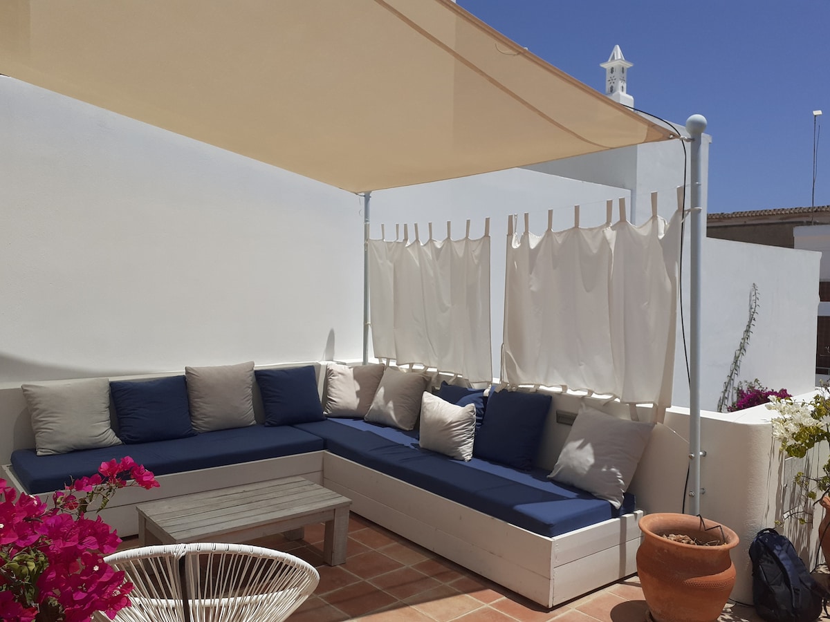 A spacious outdoor terrace features a comfortable seating area with blue cushions, complemented by soft, white drapes for privacy. Potted plants with vibrant flowers add a touch of color, while neutral-toned walls provide a serene backdrop.