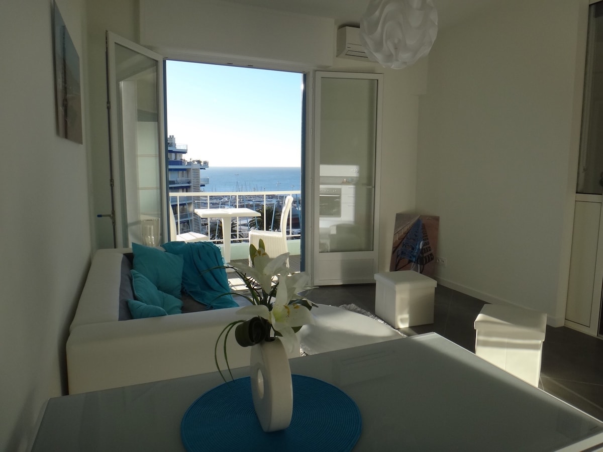 A bright and airy living space is showcased, with large glass doors opening to a view of the sea. A white sofa with colorful cushions is positioned alongside a dining table. Natural light fills the room, highlighting a decorative flower arrangement.