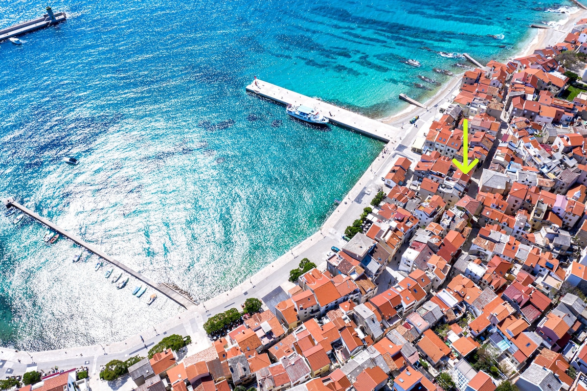 An aerial view highlights a charming village layout with terracotta roofed buildings nestled close together. The coastline features clear turquoise waters and a well-delineated promenade. A harbor with anchored boats is visible, enhancing the village's seaside character.