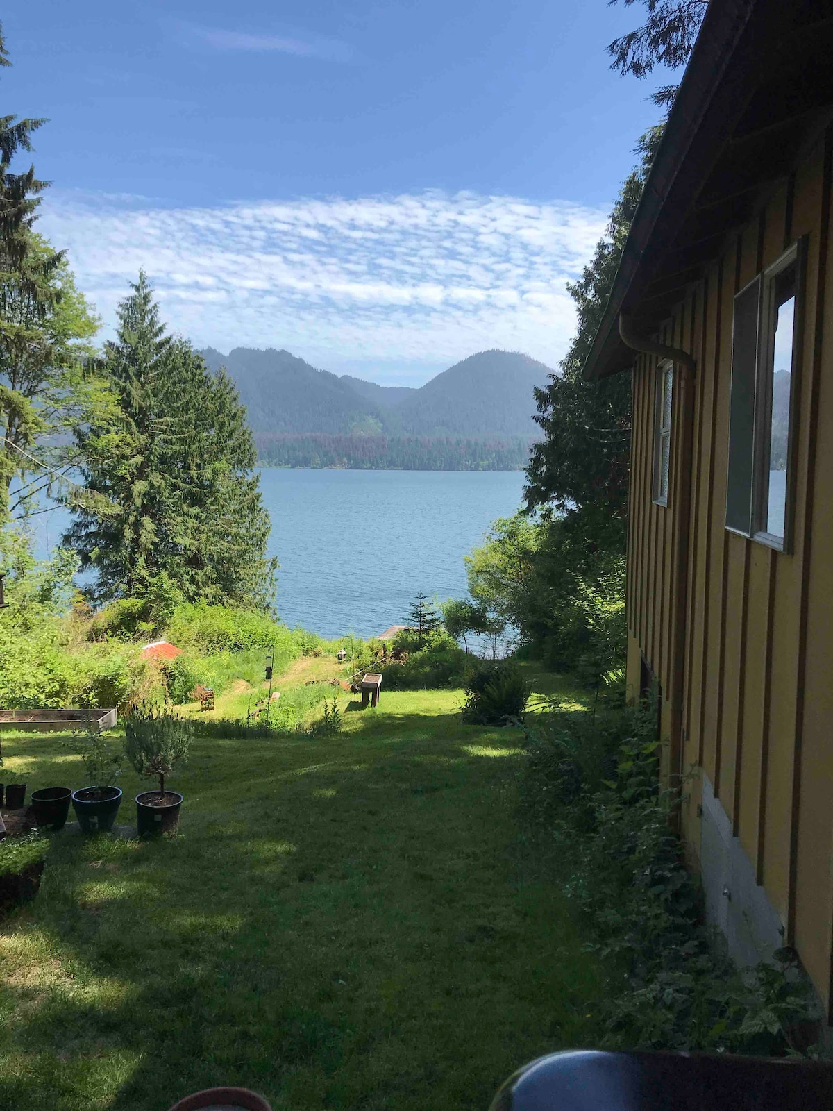 A scenic view of a lake surrounded by mountains and lush greenery is depicted. The foreground features a well-maintained lawn with several potted plants, while the yellow exterior of the house is partially visible along the right side, blending harmoniously with the natural setting.