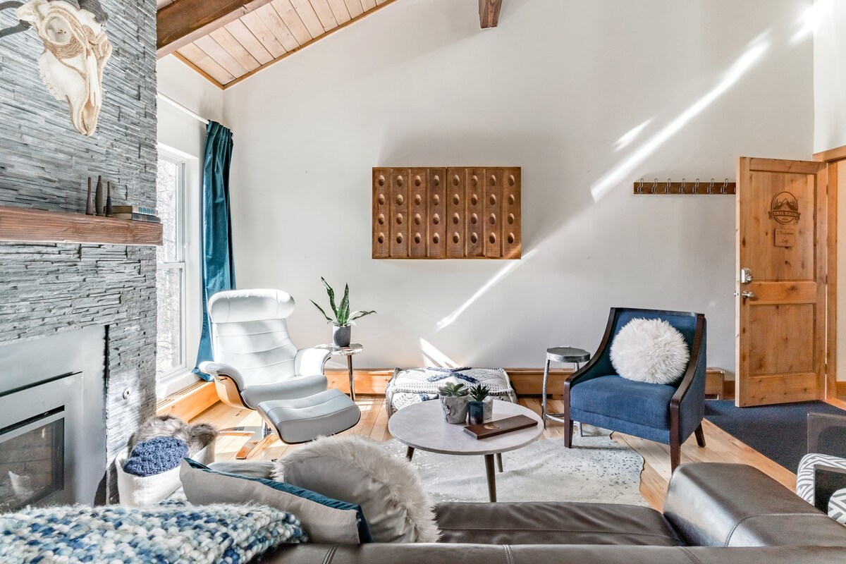 A bright living area is highlighted by a stone fireplace and wood-beam ceilings. A leather Eames chair and a blue armchair sit beside a round coffee table. Natural light filters through large windows, reflecting off the wooden accents and soft furnishings.
