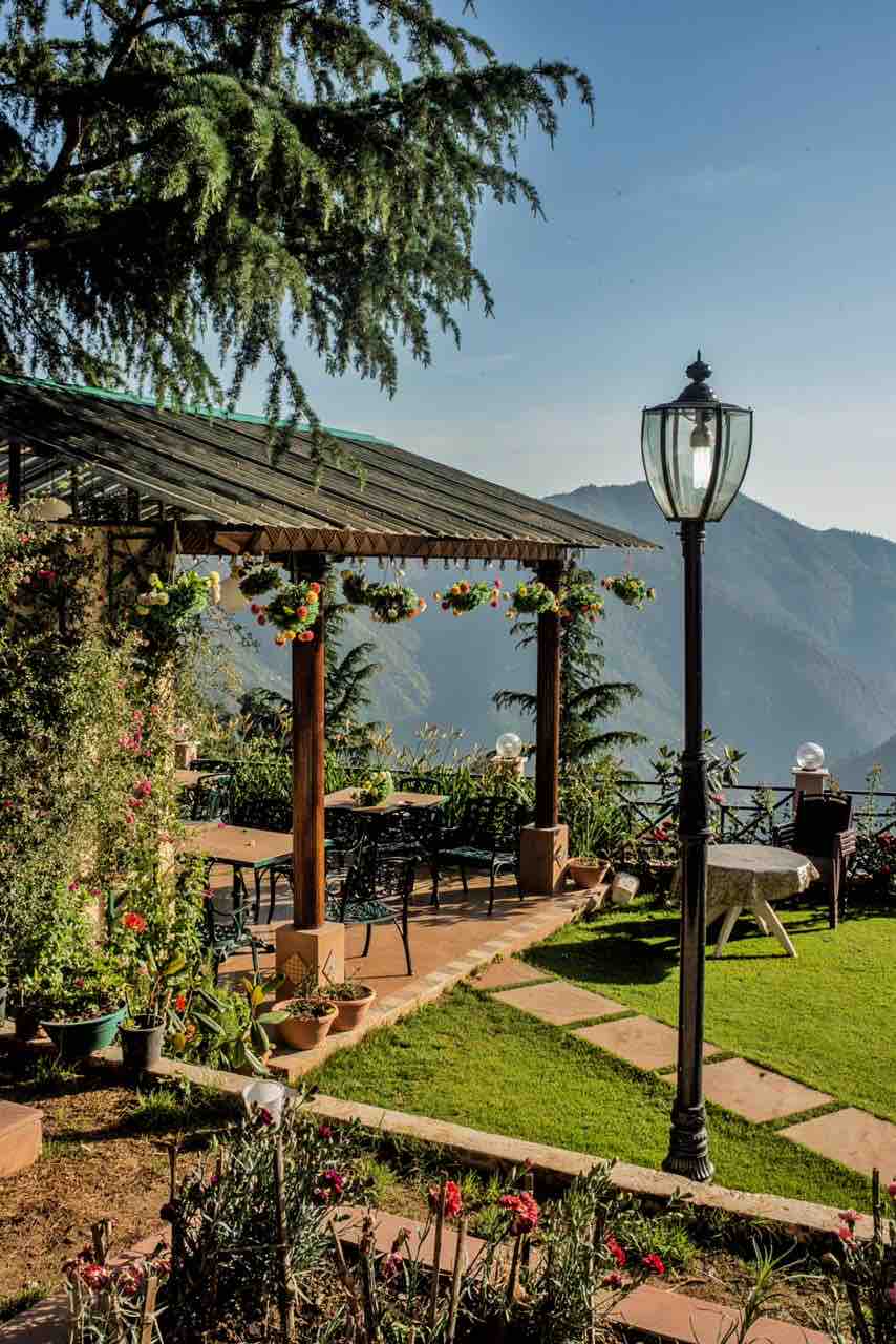 An outdoor seating area is surrounded by lush greenery and colorful flower beds. A covered wooden pergola provides shade, with tables and chairs arranged for dining. A lamp post stands nearby, offering a warm glow against the backdrop of mountain views.
