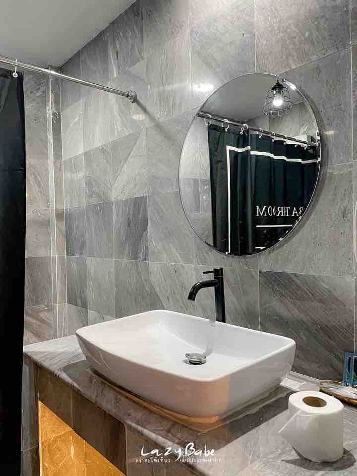 A modern bathroom is presented, featuring grey marble walls and a sleek, circular mirror above a square sink. A dark shower curtain frames the area, while a roll of toilet paper is placed on the counter, adding practicality to the elegant design.