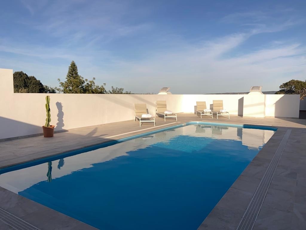 A spacious pool area is highlighted by a clear blue pool surrounded by light-colored tiles. Several lounge chairs are positioned nearby for relaxation, and a potted cactus adds a touch of greenery against a backdrop of white walls and a clear sky.