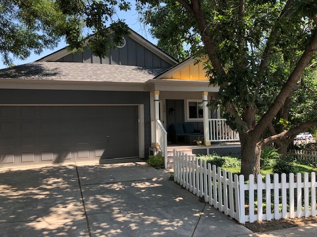 A welcoming exterior view of a home features a grey façade with a light yellow accent above the porch. A driveway leads to a garage door, and a white picket fence borders the front yard, which is complemented by lush greenery and shaded by trees.