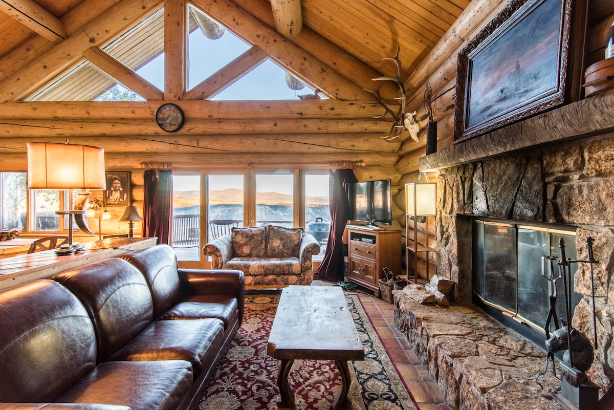 A spacious living area is highlighted by a large leather sofa and a patterned armchair, positioned near a stone fireplace. Natural light floods in through expansive windows, showcasing sweeping mountain views. Wooden beams accent the vaulted ceiling, complementing the rustic décor.