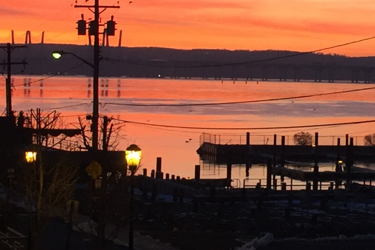 A serene sunset over the Hudson River paints the sky in warm hues, reflecting on the water's quiet surface. Silhouettes of nearby docks are visible, with lampposts softly illuminating the scene. Electric lines and poles frame the image, adding to the tranquil riverside atmosphere.