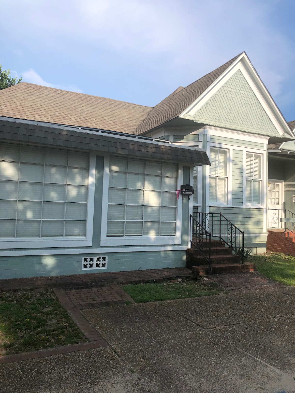 The exterior of a historic building features a light blue façade with large windows covered by white blinds. A set of steps leads to the entrance, framed by the intricate gables of the roof. Brick pavers and grass surround the property, providing a welcoming appearance.