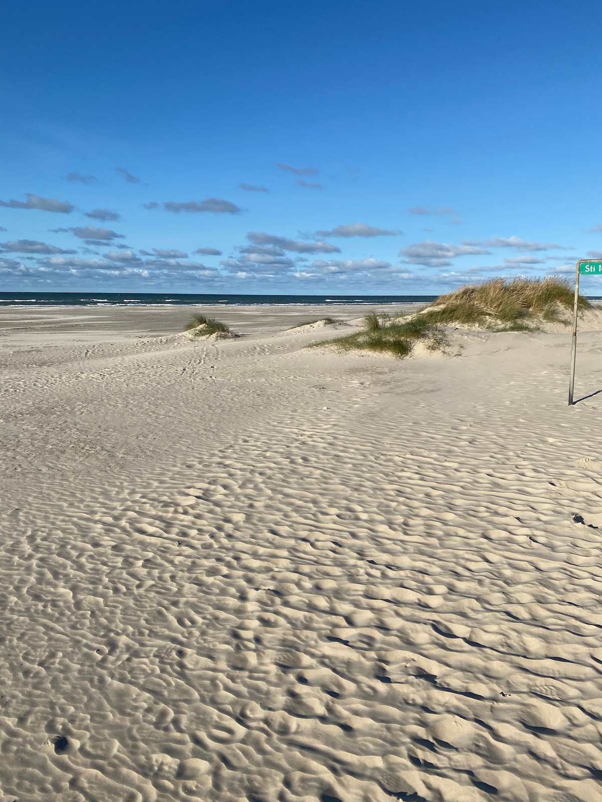 The sandy beach extends beneath a clear blue sky. Gentle dunes are visible in the distance, with grasses swaying softly in the breeze. The shore meets the calm sea, where small waves create a tranquil atmosphere. Scattered clouds contribute to a serene coastal scene.