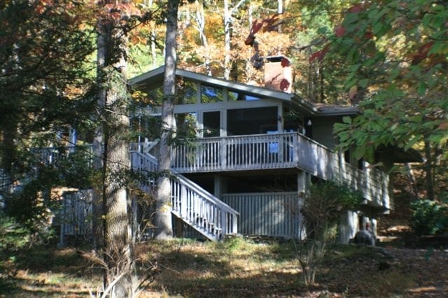A modern lakefront home is nestled among trees, showcasing a spacious deck and stairs leading to the entrance. Large windows allow for natural light, while the surrounding autumn foliage adds vibrant color to the serene setting.