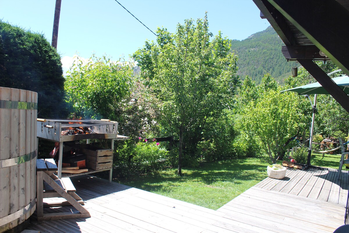 An outdoor terrace is visible, featuring a barbecue area and wooden deck. Lush greenery and trees create a natural setting, while a shaded area is provided by an umbrella. A glimpse of mountains can be seen in the background, adding to the serene atmosphere.
