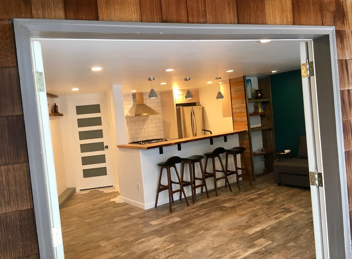An open-plan space is showcased, featuring a kitchen area with stainless steel appliances and a bar-style countertop with tall stools. The walls are adorned in warm wood tones and a rich green accent. Soft lighting provides a cozy ambiance.