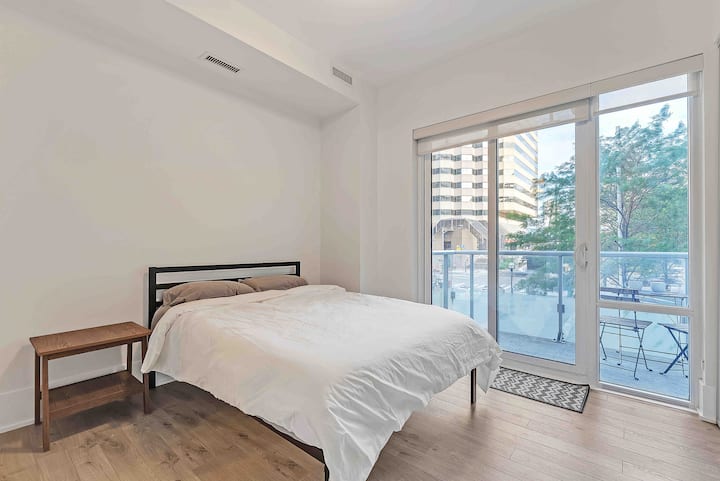 غرفة نوم مشرقة وجيدة التهوية مع سرير مريح بحجم كوين، وبياضات نظيفة، ونوافذ ممتدة من الأرض إلى السقف تؤدي إلى شرفة خاصة - ملاذك المريح في قلب المدينة.