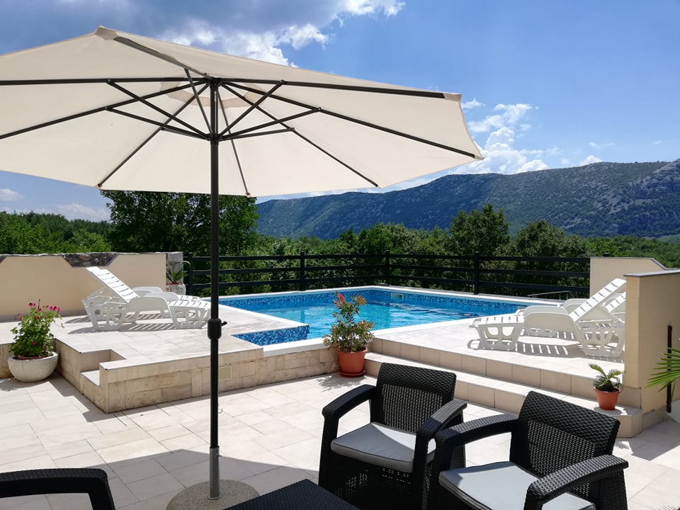A spacious patio area features a large umbrella providing shade over seating arrangements. In the background, a clear blue swimming pool is bordered by landscaping, with green mountains visible beyond. Sun loungers are positioned around the pool, enhancing the area’s inviting atmosphere.