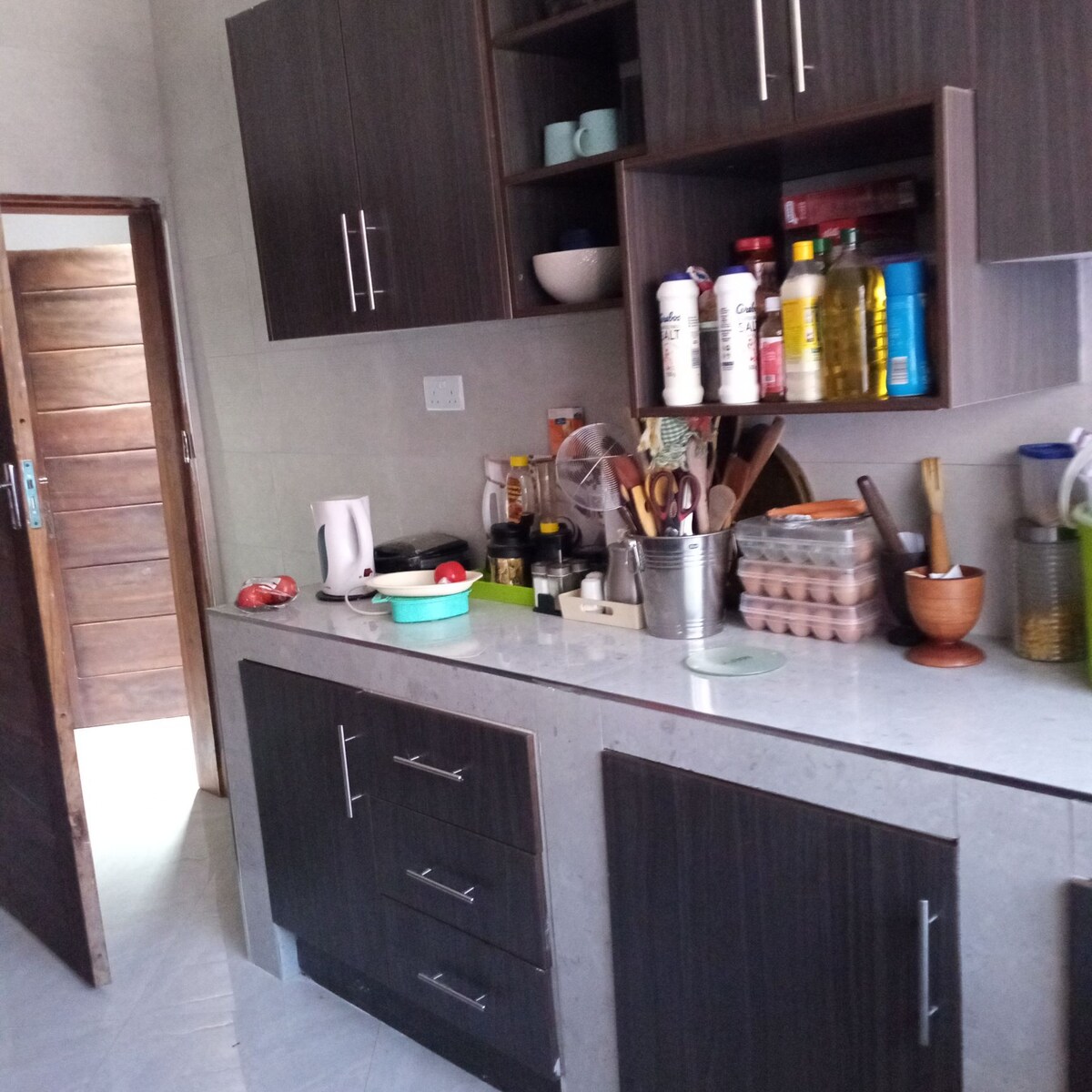 A contemporary kitchen features dark wood cabinets with various supplies neatly organized on a countertop. Essential appliances, such as a kettle and stovetop, are present. A small window illuminates the space, while a wooden door leads to another area of the home.