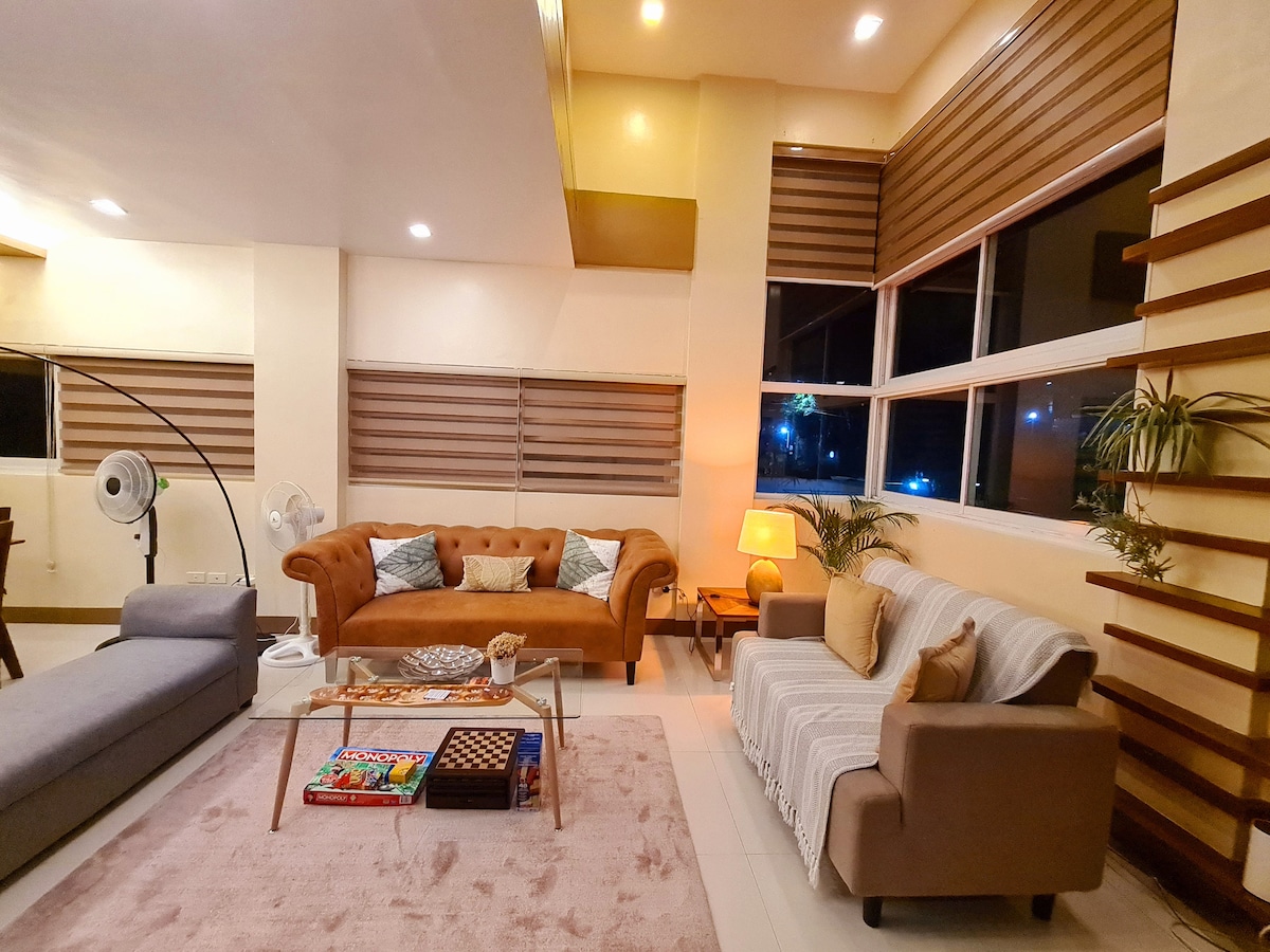 The living area features a blend of modern and cozy furnishings, including a brown sofa, a light gray lounge chair, and a striped throw blanket draped over a second couch. Large windows allow natural light to brighten the space, complemented by soft lighting from a floor lamp.