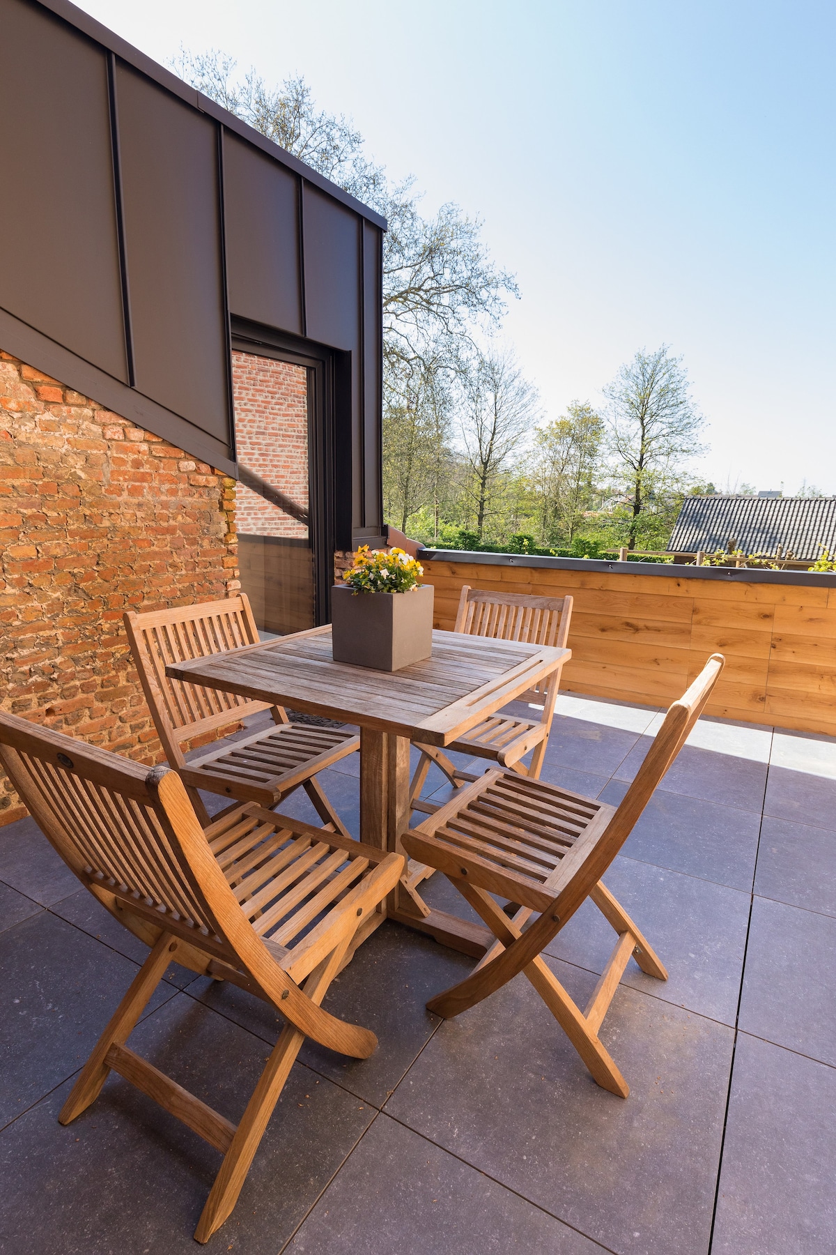 A wooden outdoor dining set is arranged on a terrace. The set includes a square table and four foldable chairs, all crafted from natural wood. A planter with blooming flowers sits at the center of the table. The space is surrounded by a wooden railing and offers views of greenery.