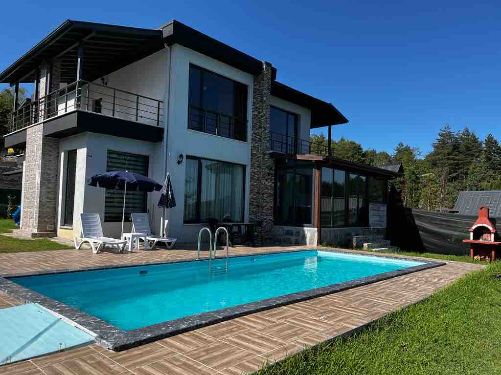 The exterior of a modern house features a spacious outdoor area with a swimming pool surrounded by sun loungers. A patio umbrella provides shade nearby. Lush greenery and trees border the property, complementing the home's sleek design and large windows.