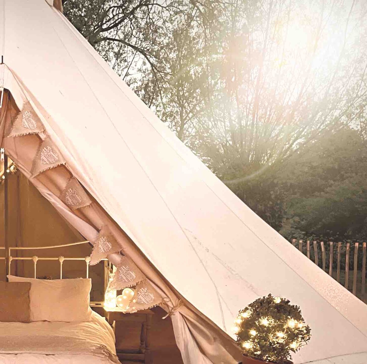 A cozy bell tent is illuminated by soft lighting, showcasing a spacious bed with neutral bedding. The canvas structure features decorative trim and is framed by natural greenery, creating a tranquil setting surrounded by nature.