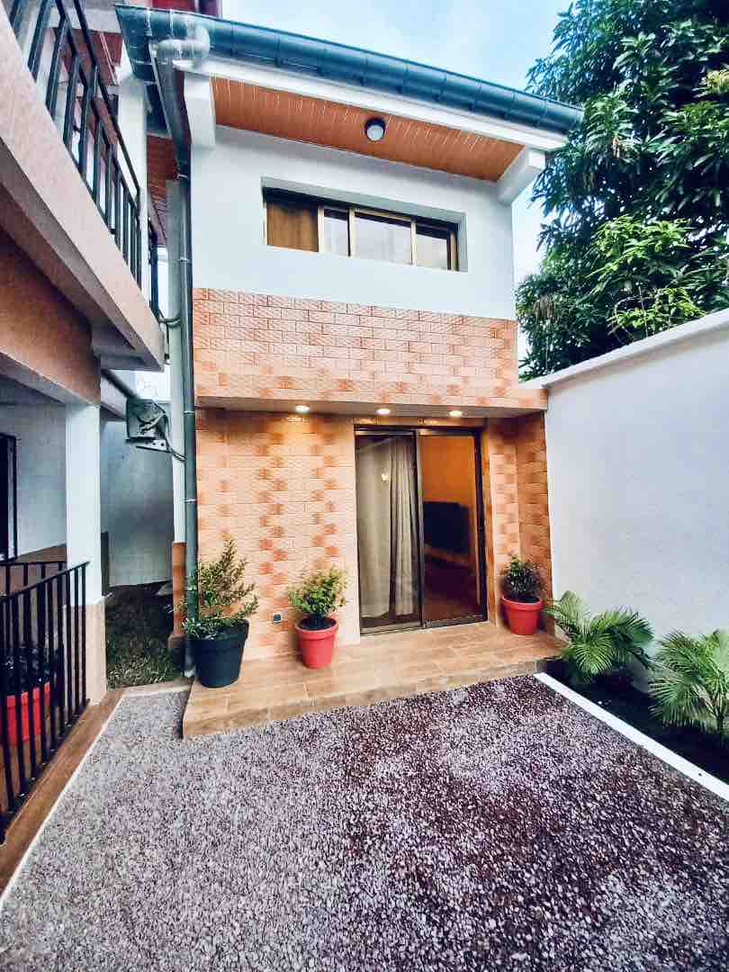 An entranceway is visible, featuring a decorative brick façade and lush greenery lining the path. Potted plants are situated on either side of the entry door, which leads to a well-lit interior space. The surrounding area is clean and organized, with a gravel surface for easy navigation.