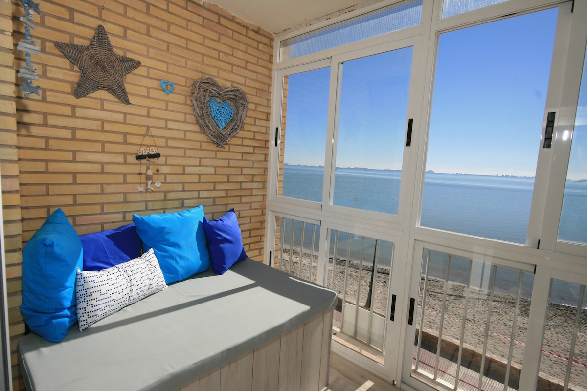 A cozy lounge bed is positioned against a wall of light brick, adorned with decorative nautical items. Bright blue cushions complement the neutral bedding. Glazed windows frame a panoramic view of the serene beach and water beyond, allowing natural light to fill the space.