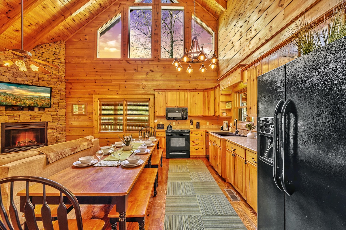 An open-concept kitchen and dining area features a large wooden table set for a meal. Light from large windows fills the space, illuminating the wood-paneled walls and stone fireplace. A black refrigerator stands alongside the kitchen appliances, creating a functional cooking space.