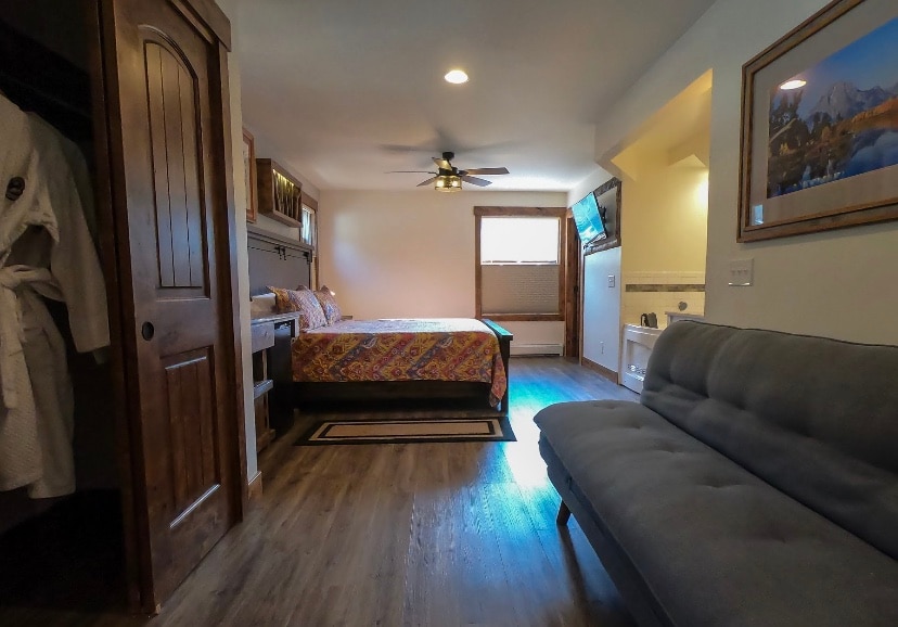 A bright and spacious room features a colorful patterned bedspread covering a large bed. A comfortable gray couch sits at the foot of the bed. Light from a window illuminates the wooden floors, while a ceiling fan adds a functional touch to the space.