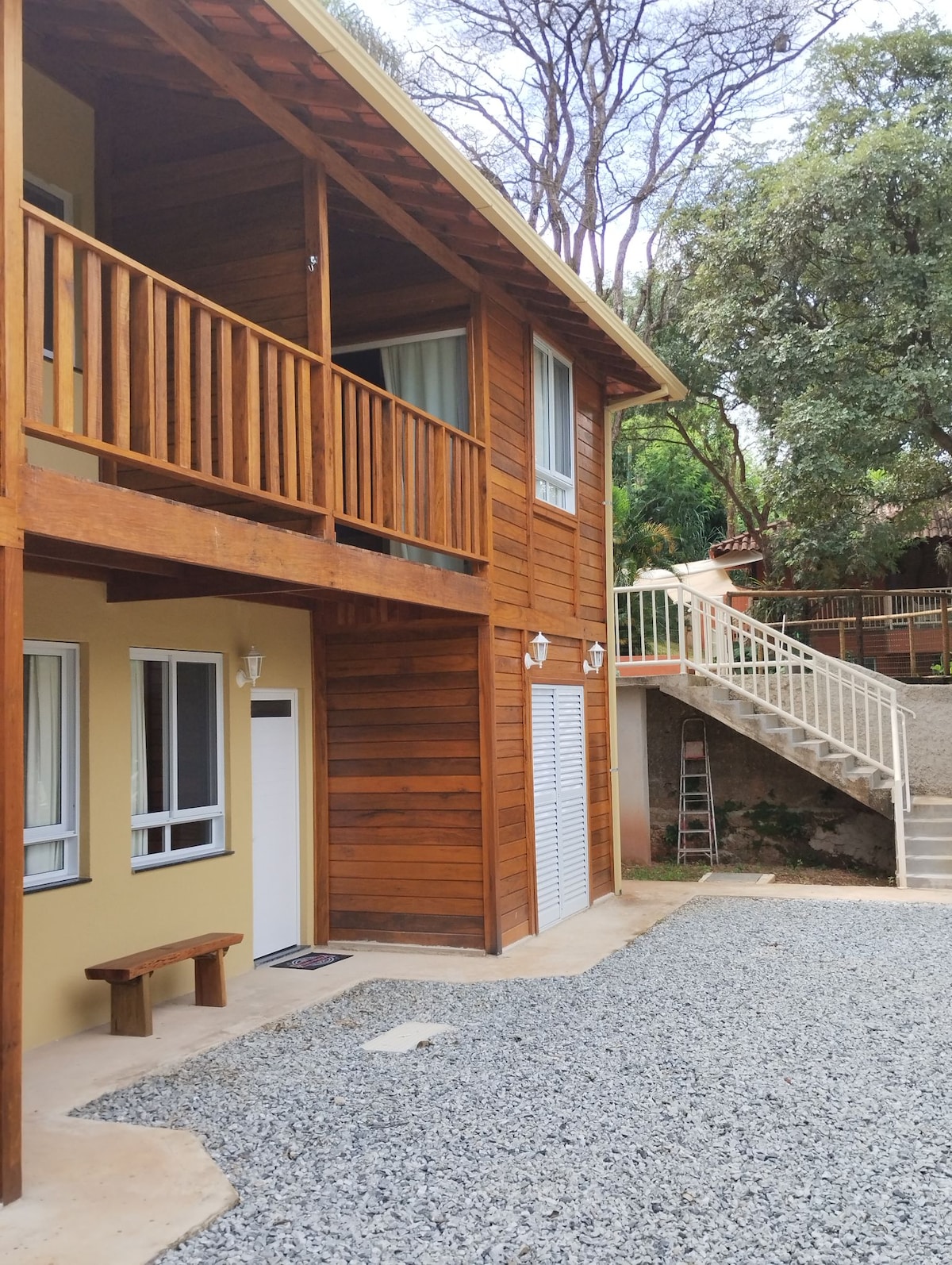 A two-story wooden structure with a rustic design is shown, featuring a wrap-around balcony. The lower level includes a bench and an entrance door, with light-colored walls contrasting against the wooden exterior. Gravel pathways and greenery surround the building, adding to the serene environment.