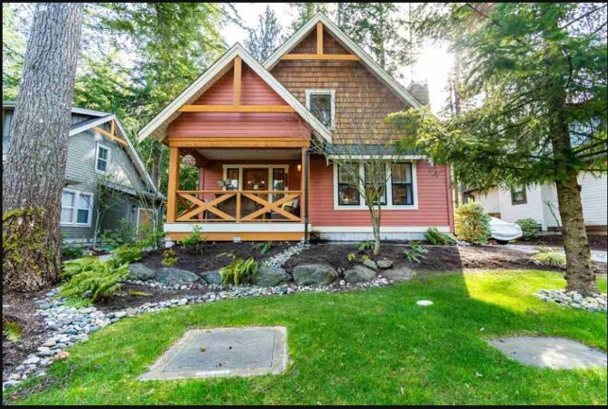 The charming craftsman cottage features a welcoming porch and vibrant exterior, surrounded by lush greenery. A well-maintained lawn with rock accents enhances the entrance, while the surrounding tall trees provide a serene natural backdrop.