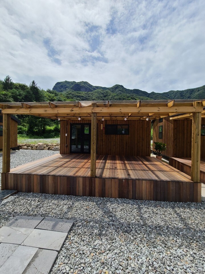 A wooden structure is presented on a landscaped area featuring a spacious deck. Natural stone and gravel surface create a rustic feel. The background features a mountain landscape under a partly cloudy sky, adding a natural setting to the glamping site.