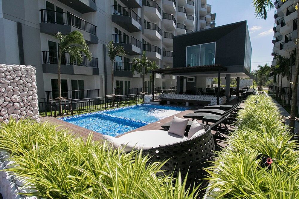 A swimming pool is framed by lush green landscaping and lounge chairs. Modern condominium buildings are visible in the background, with a shaded area featuring seating near the water. Sunlight reflects off the pool's surface, creating a serene and inviting environment.