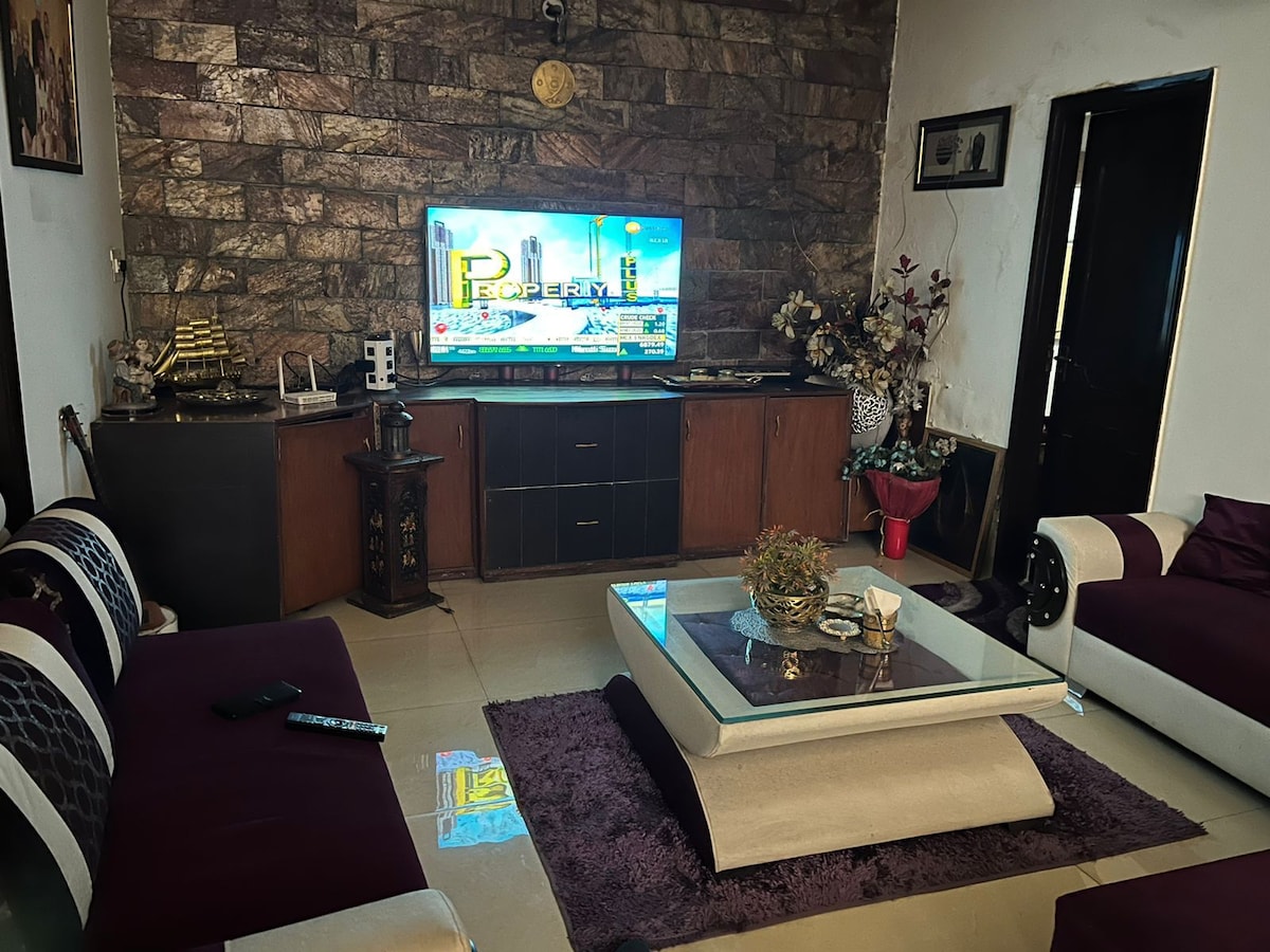 The living area features a contemporary design with stone wall accents and a large television mounted above a dark wood media unit. A glass-topped coffee table sits on a plush purple rug, surrounded by comfortable seating in contrasting colors.