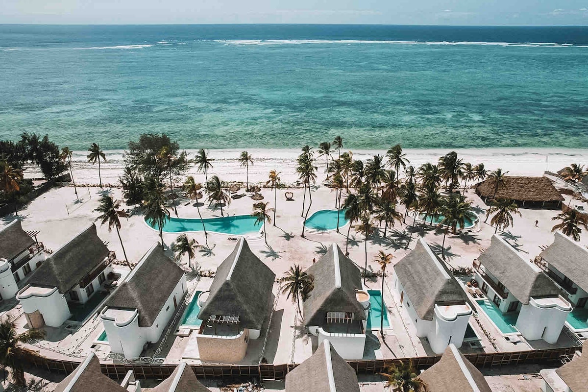 An aerial view captures a serene beachfront scene with white structures featuring thatched roofs. Lush palm trees surround three clear swimming pools, which are linked by pathways. The vibrant blue ocean extends into the distance, complementing the sandy beach in the foreground.