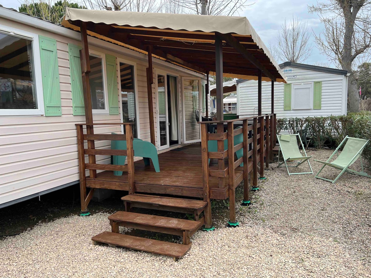 A wooden deck is featured at the entrance of the mobilhome, with steps leading up to a covered area. Green chairs are positioned nearby, and the exterior showcases light-colored siding and green shutters. Surrounding shrubbery adds a natural touch.