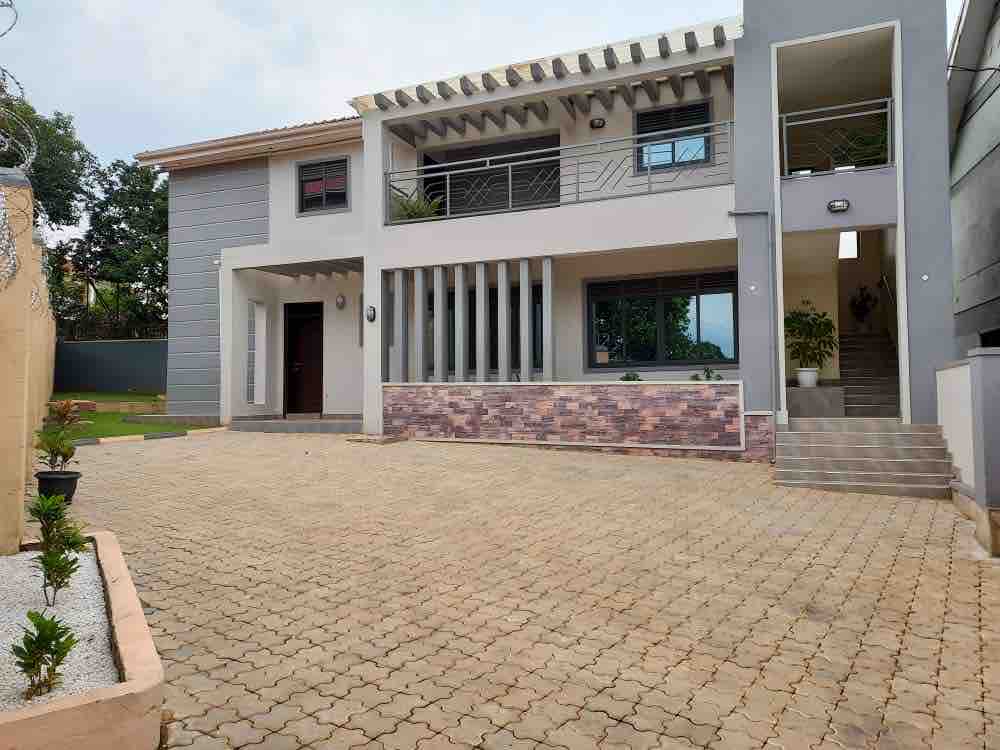 The modern two-story building features a spacious entrance with a patterned stone pathway. Large windows are present on both levels, allowing natural light to fill the interior. Lush greenery is visible in the surrounding area, enhancing the tranquil setting.