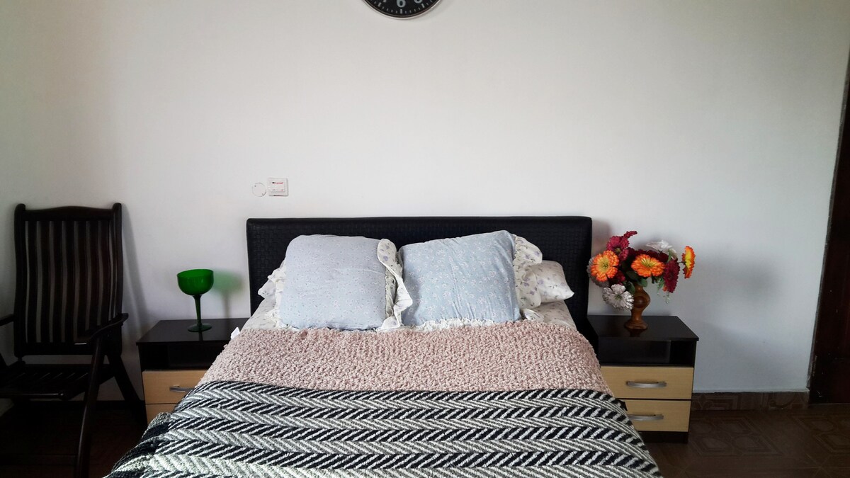 A cozy bedroom is depicted with a spacious bed adorned with soft pillows and a knitted blanket. A dark wood rocking chair is positioned nearby, and nightstands feature small lamps and decorative flowers, contributing to a relaxed ambiance.