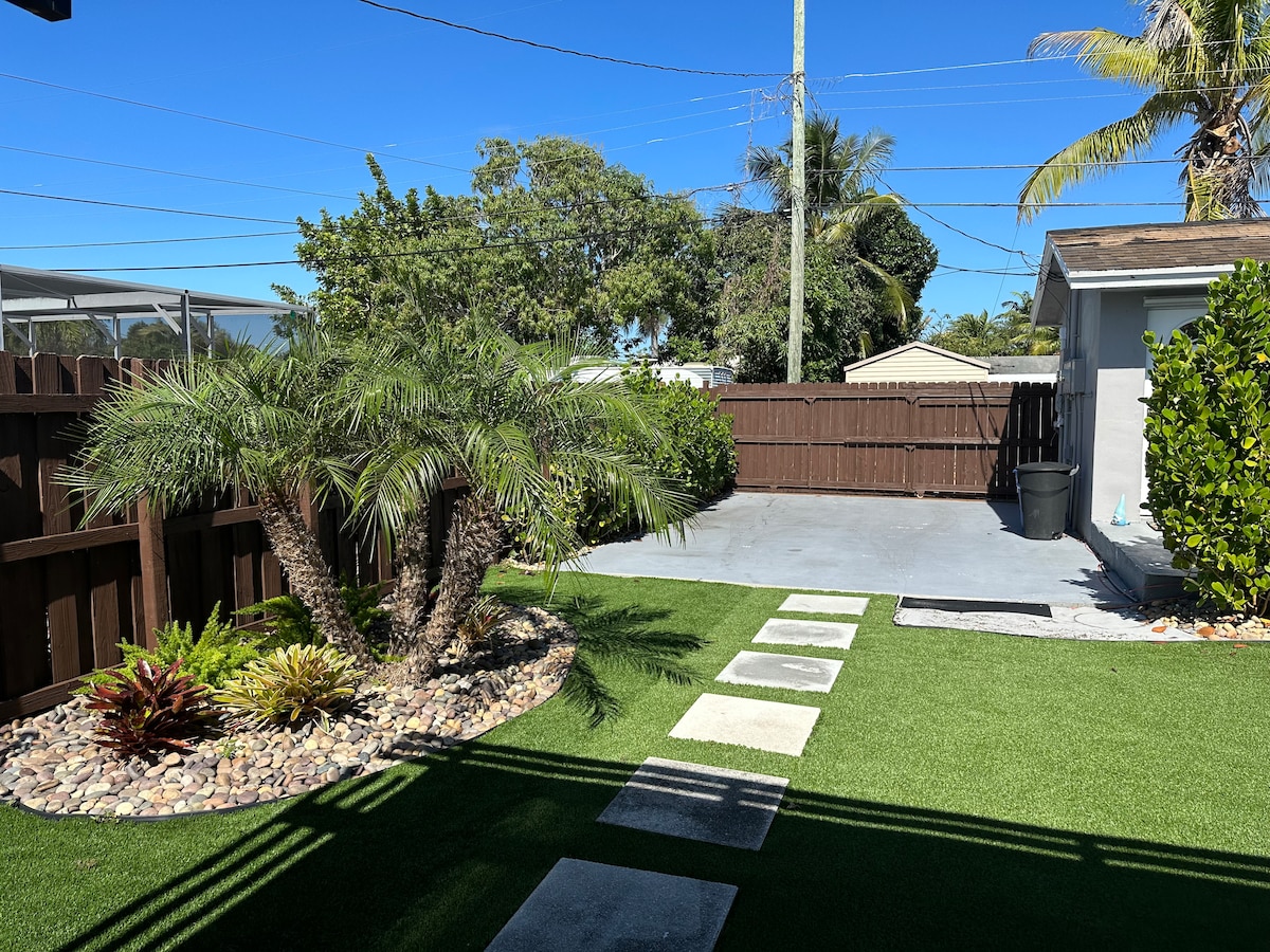 A landscaped outdoor space features a palm tree and low-maintenance plants, with stone pathways leading through a lush green lawn. A fenced area and a paved section are visible, providing a serene environment for relaxation.