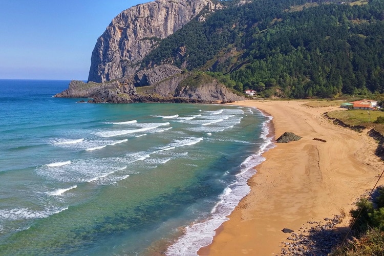 A serene beach scene is depicted, showcasing gentle waves lapping against sandy shores. Rolling hills covered in greenery rise in the background, with rocky cliffs adding texture to the landscape. The clear blue waters and sandy beach invite relaxation and tranquility.